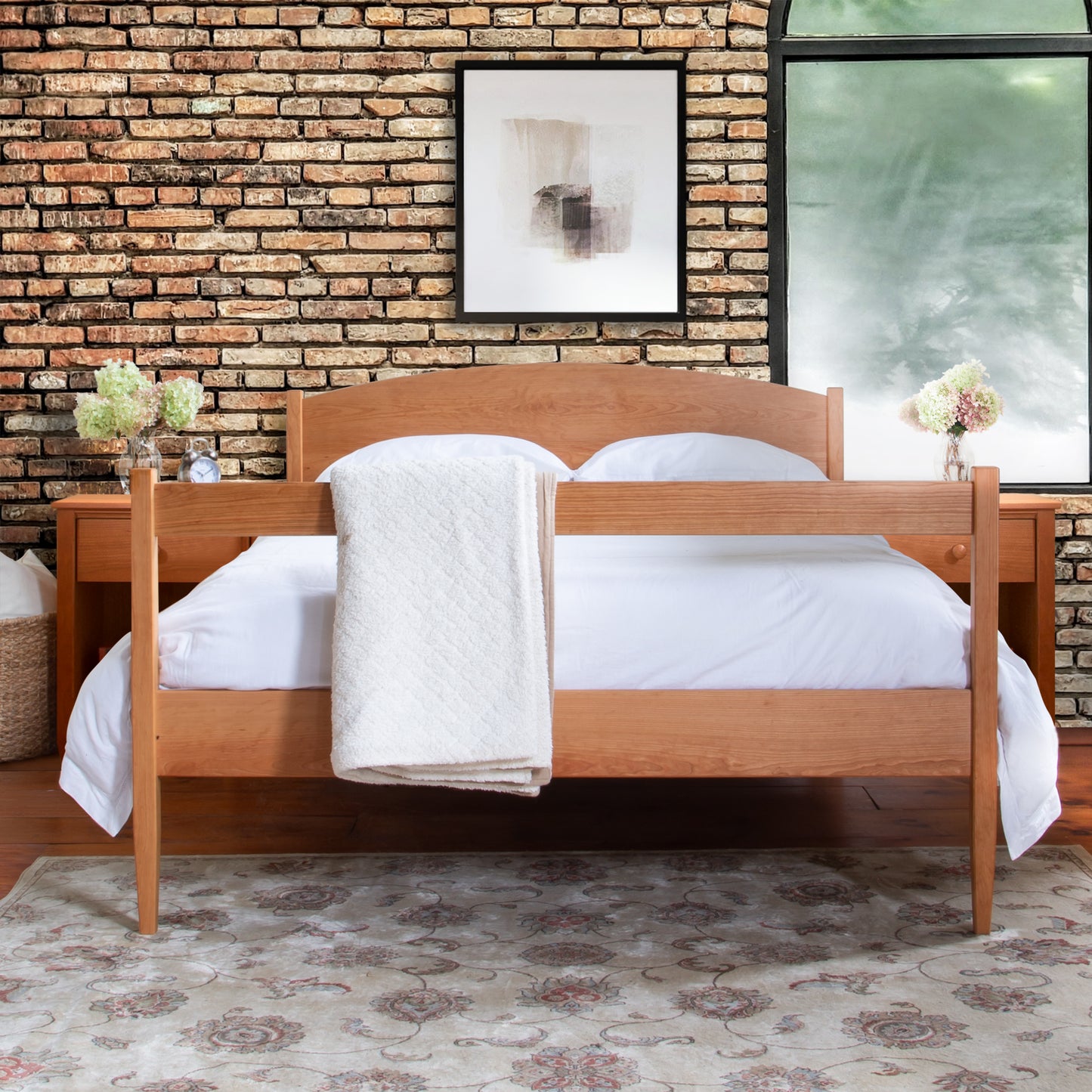 A Maple Corner Woodworks Vermont Shaker Bed made of wood, with white bedding and a white throw blanket, flanked by two small nightstands with flowers, placed in a room with a brick wall and a framed.