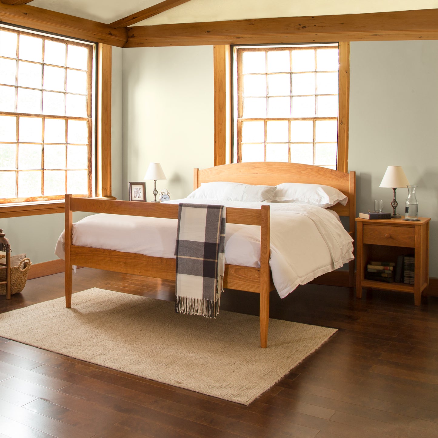 A neatly made Maple Corner Woodworks Vermont Shaker Platform Bed with white bedding and a plaid throw blanket in a room with wooden floors, rustic framed windows, and matching wooden nightstands with lamps.
