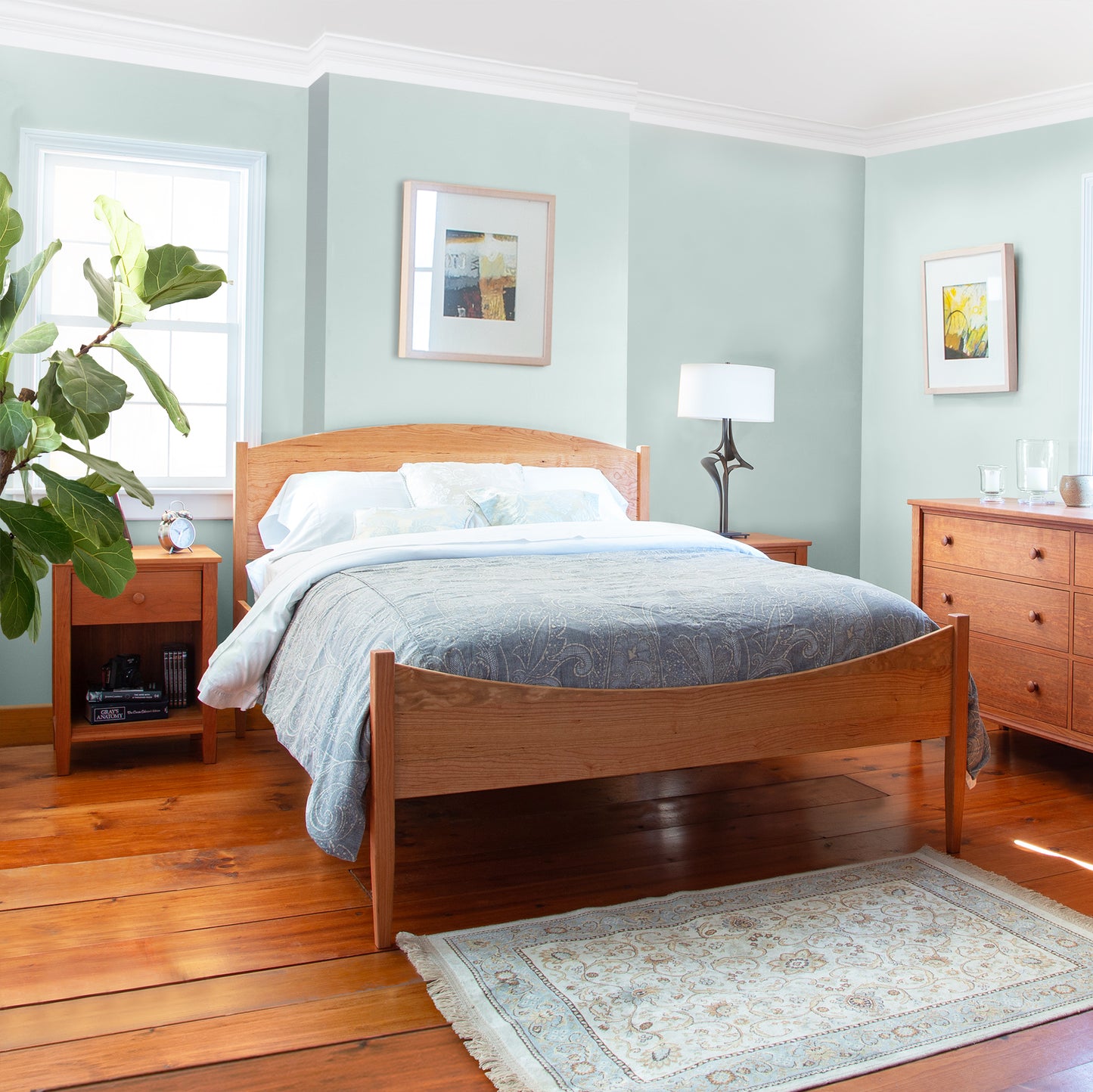 A neatly presented bedroom with a Vermont Shaker Moon platform bed, matching dresser, and side table from the Maple Corner Woodworks Vermont Shaker Furniture Collection, set against a light green wall with framed artwork.
