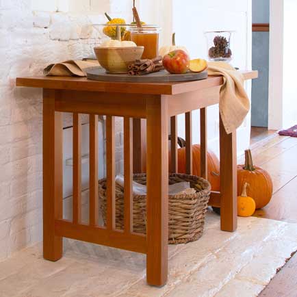 A wooden table against a white brick wall holds a bowl of apples, a jar of honey, a wooden cutting board, and a napkin. Below the table, there is a wicker basket with rolls. Pumpkins are placed on the floor to the right of the table.