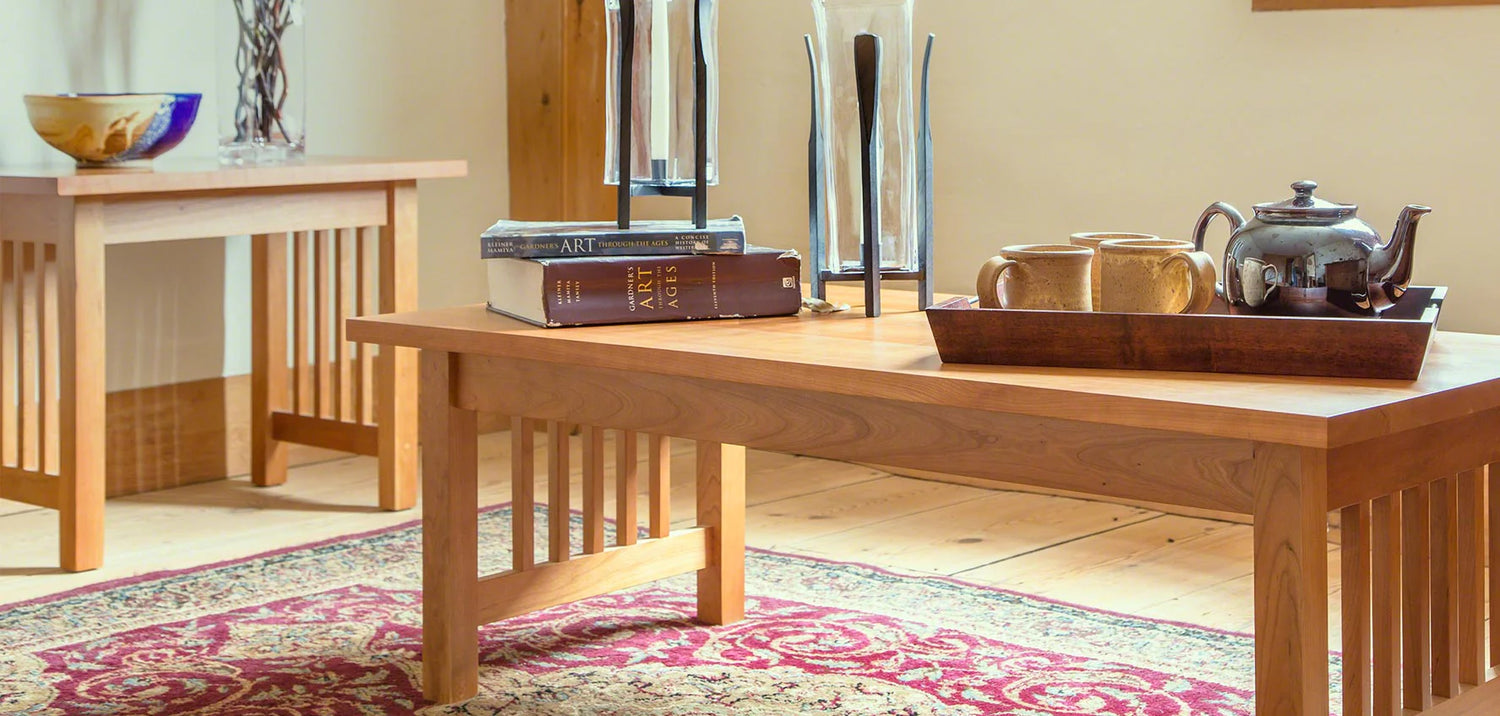 A cozy living room features two wooden tables with slatted sides. The foreground table holds books, a tea set, and a decorative tray, while the background table displays a bowl. Both tables rest atop a red patterned rug on a wooden floor.