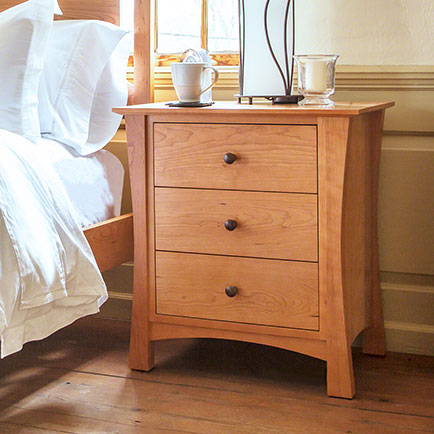A wooden bedside table with three drawers, each with a round knob, sits beside a bed. On top of the table are a white mug, a glass of water, and a small lamp with a rectangular shade. The scene is lit by daylight coming through a nearby window.