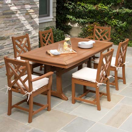 Outdoor dining set with wooden table and six chairs on a patio. The table has a pitcher of lemonade and a stack of plates. Each chair has a white cushion tied to it. The patio is paved with large stones, and greenery is visible in the background.