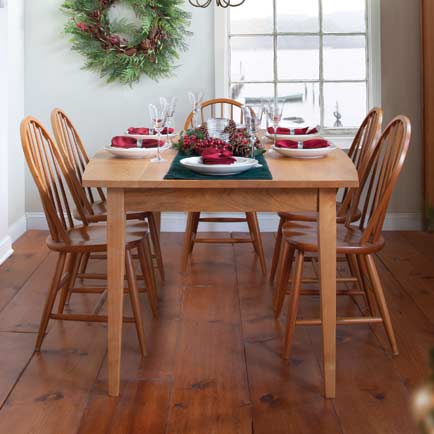 A wooden dining table is set for six in a dining room. Red napkins and white plates are placed at each seat, and a festive centerpiece with candles and greenery is in the middle. A wreath hangs on the wall, and a window shows a snowy outdoor scene.