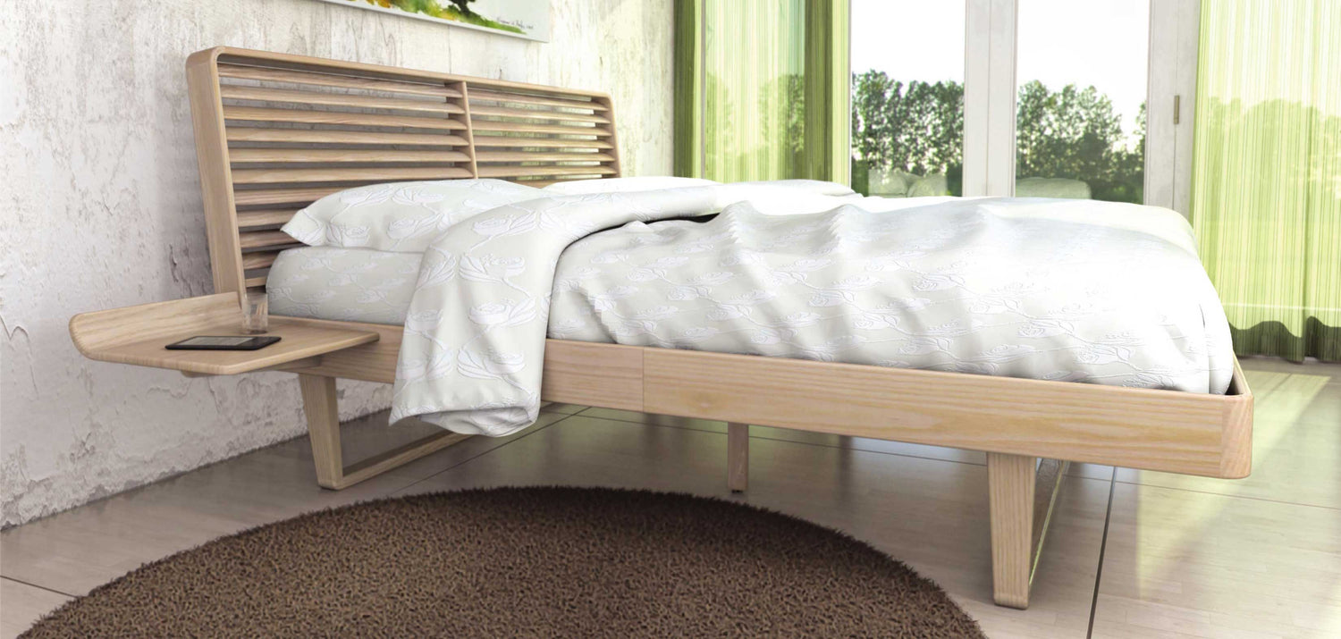 A modern wooden bed with a slatted headboard and a small attached side table holding a glass and a phone. The bed has white bedding with a subtle floral pattern. The room has green curtains and large windows showing greenery outside. A round brown rug is on the floor.