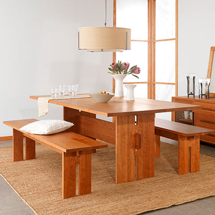 A dining room features a wooden table with a light brown finish, accompanied by two matching benches. The table is set with glassware, a bowl, and a vase with flowers. A modern pendant light hangs above, and a light brown rug covers the floor.