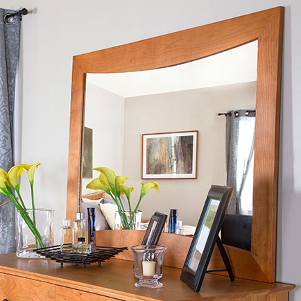 A wooden-framed mirror hangs above a wooden dresser. On the dresser are two vases with yellow flowers, a lit candle, several bottles, and a framed photo. Reflections of light gray walls and curtains are visible in the mirror.