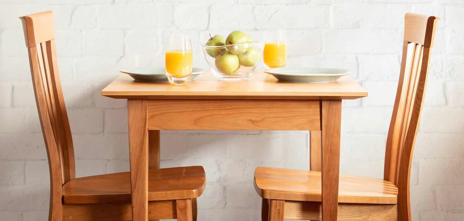 A small wooden dining table with two matching wooden chairs is set against a white brick wall. The table has two plates, two glasses of orange juice, and a clear bowl containing green apples.