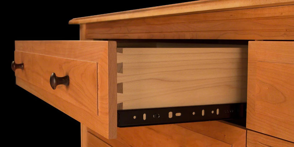 A close-up view of a wooden drawer partially open, showcasing the dovetail joints and the metal slide mechanism. The drawer is part of a light-colored wooden cabinet with dark-colored knobs. The background is completely black.