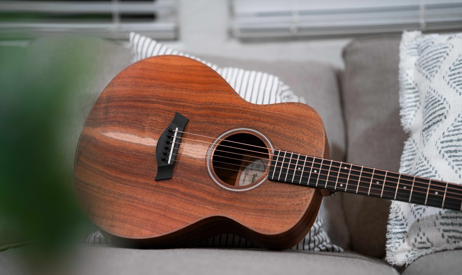 An acoustic guitar with a wooden finish is resting on a gray couch, partially leaning against white and striped cushions. The background includes a blurred-out window with closed blinds.