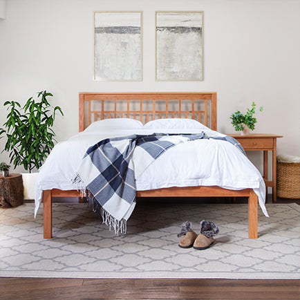 A bedroom features a wooden bed frame with white bedding and a checkered throw blanket. Two abstract paintings hang above the headboard. A potted plant and a wooden nightstand with a plant sit beside the bed. Slippers are placed on a patterned rug.