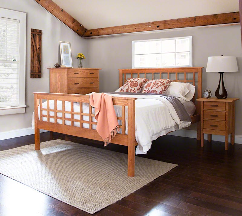 A bedroom with wooden furniture, including a bed with a gridded headboard and footboard, a nightstand with a lamp, and a dresser. The bed is made with white bedding and decorative pillows. There is a window on the back wall and a rug on the polished dark floor.