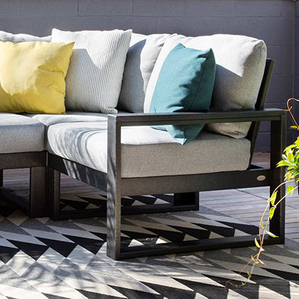 Outdoor sectional couch with light gray cushions and three pillows in yellow, teal, and white with stripes. The couch has a black metal frame and sits on a geometric patterned rug on a wooden deck. Some green plants are visible in the corner.