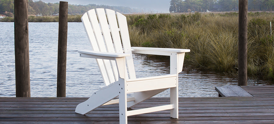 A white Adirondack chair is placed on a wooden dock overlooking a body of water. Tall grasses and trees can be seen in the background. The scene is calm and serene.