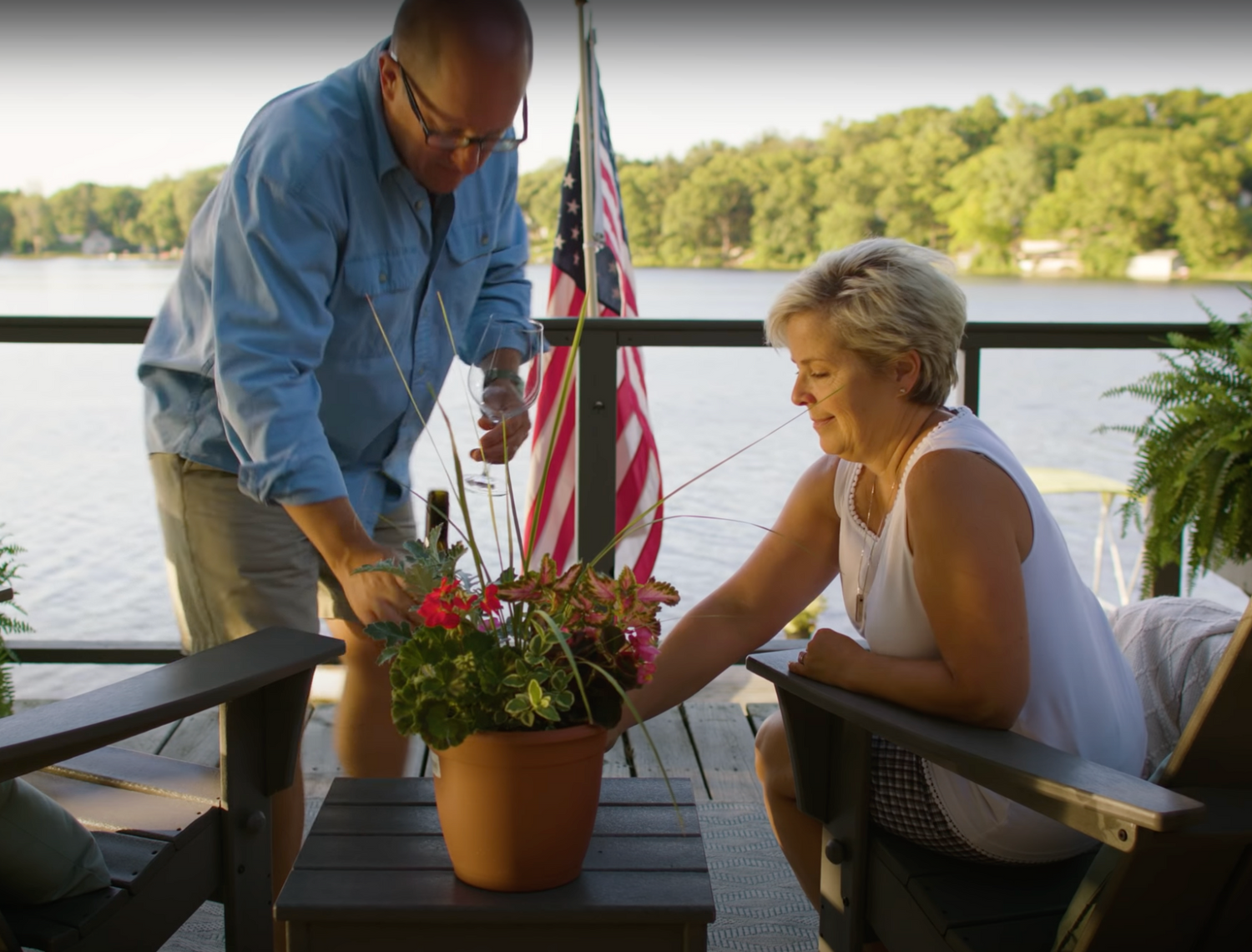 Two people are tending to a potted plant on a lakeside patio. The man is watering the plant while the woman adjusts it. They are outdoors with a view of a lake and trees in the background. An American flag is displayed in the background.
