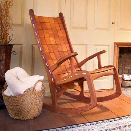 A wooden rocking chair with a woven seat and backrest is placed in front of a paneled wall. Beside it, there is a wicker basket filled with white blankets. The floor is wooden, and part of a rug and fireplace are visible in the background.