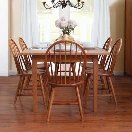 A wooden dining table is set with six place settings, including plates, bowls, and cups. Six wooden chairs surround the table, and a bouquet of flowers is in the center. The room features hardwood floors, white curtains, and a chandelier above the table.