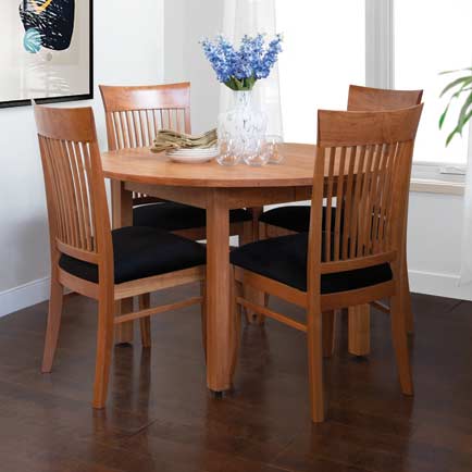 A round wooden dining table with four matching wooden chairs with black cushions. A vase of blue flowers, a plate, and several glasses are on the table. The room has a window with white curtains and a framed artwork on the wall.