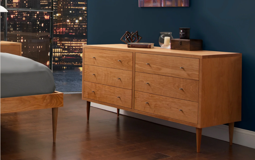 A wooden dresser with several drawers stands against a dark blue wall in a bedroom with a cityscape visible through the window at night.
