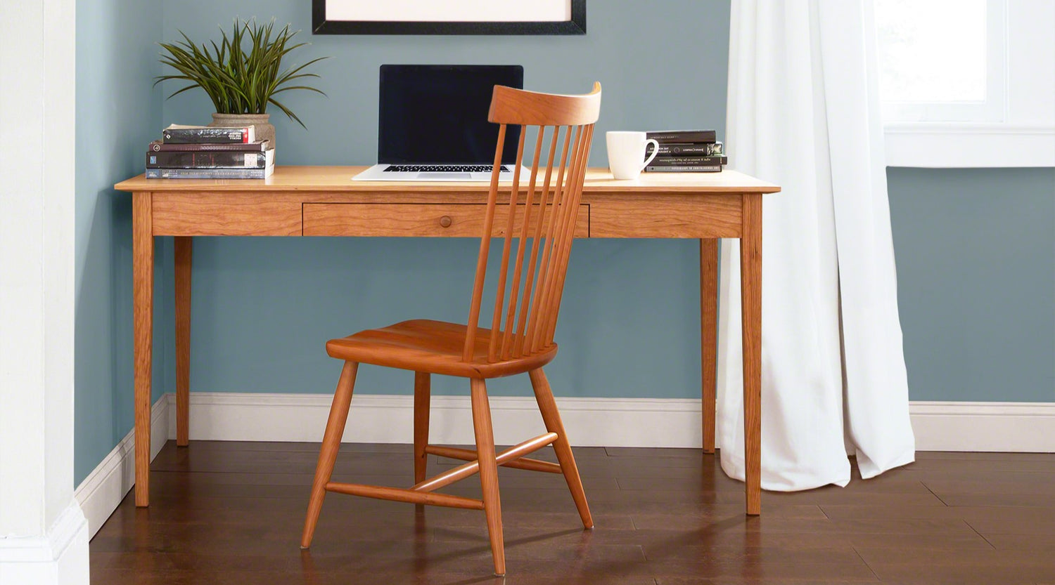 A wooden desk with a laptop and books on it, paired with a matching chair in a room with blue walls and dark flooring.