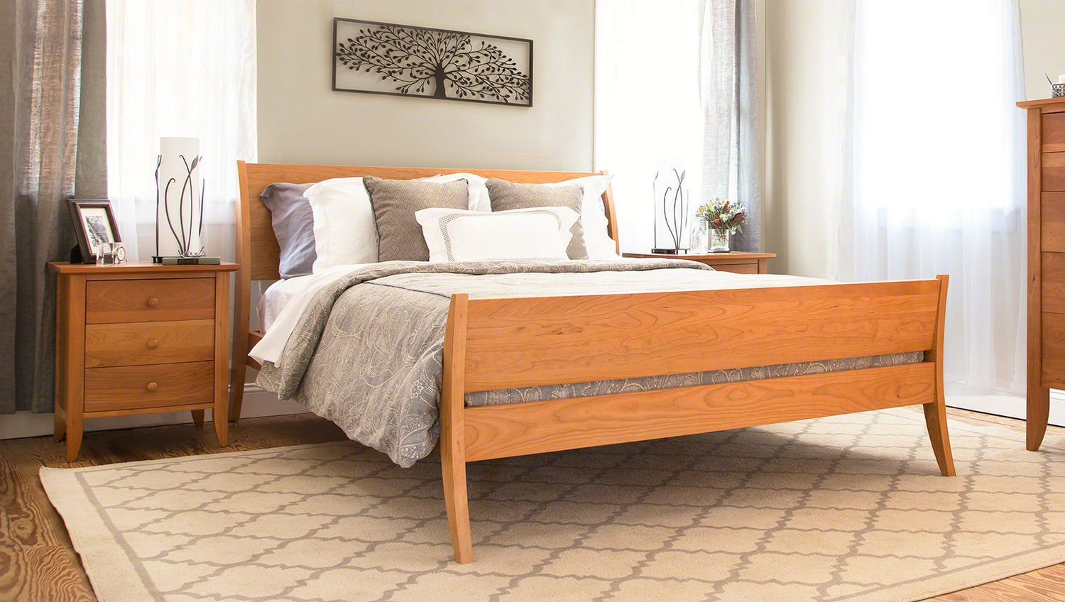 A neatly arranged bedroom featuring a wooden bed with white and beige bedding, two matching wooden nightstands with lamps, and a patterned beige rug on a hardwood floor. light curtains hang by the window.