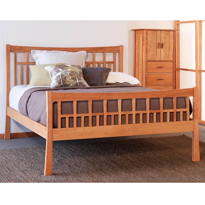 A wooden bed with a slatted headboard and footboard, topped with a quilt, pillows, and a throw blanket. A matching wooden dresser with drawers and a cabinet is positioned to the side in the background.