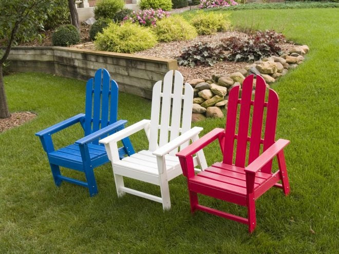 Three brightly colored adirondack chairs (blue, white, and red) arranged on a lush green lawn, surrounded by a well-manicured garden and stone border.