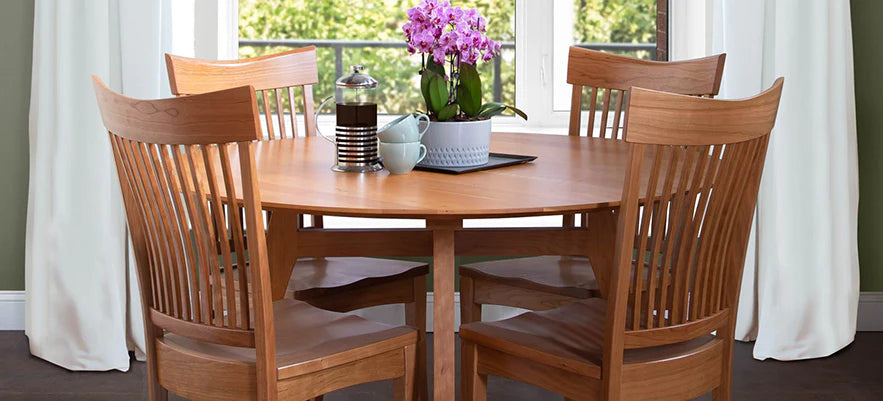 A wooden dining table set with four chairs, a centerpiece of purple flowers, and a coffee pot with cups on a placemat, positioned in front of a large window with white curtains.