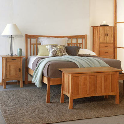 A bedroom with a wooden bed frame, a nightstand, and a storage bench at the foot of the bed. The bed is made with brown and gray bedding, and a light blue throw blanket. A table lamp sits on the nightstand, and a wardrobe cabinet stands against the wall.