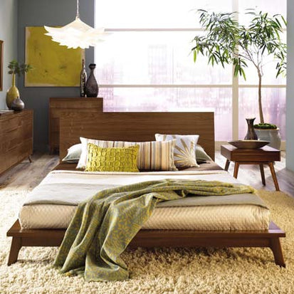 Wooden bed with green and beige bedding, decorative pillows, dresser, and side table in natural lighting.