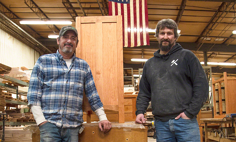 Two men showcasing handcrafted wooden furniture in a woodworking shop.