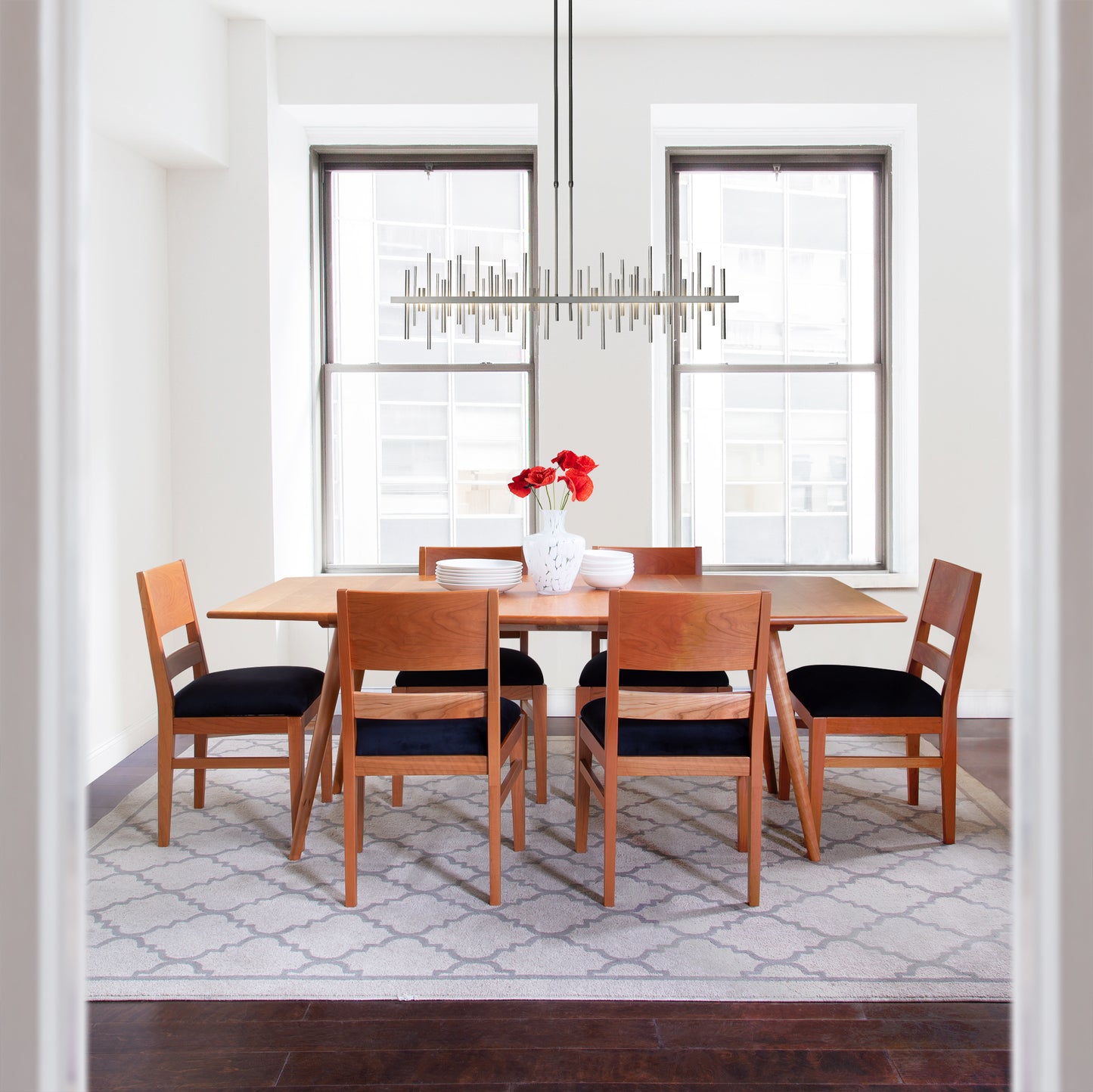 A modern dining room with a natural solid wood table, six Vermont Woods Studios Burke Modern Chairs, and a geometric chandelier, with natural light coming through two windows.