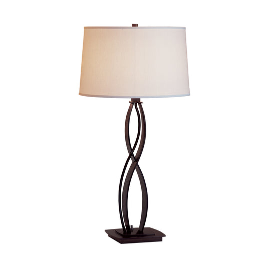 A Hubbardton Forge Almost Infinity Table Lamp with an intertwined wrought iron base design and an oval-shaped white shade, isolated on a white background.