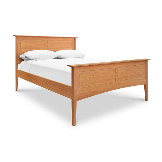 A handmade wood bed from Maple Corner Woodworks, known as the American Shaker Panel Bed, is showcased here with a natural finish and simple design. This bed features a headboard and footboard with panel details and is made from sustainably harvested woods. It sports white bedding and two pillows, while clean lines and tapered legs complete the look.