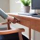 A person is pulling open a drawer of an American Shaker Writing Desk by Maple Corner Woodworks, made from sustainably harvested hardwood by skilled Vermont craftsmen. On top of the desk sits a keyboard and computer monitor, alongside a pot with a green plant. In the foreground, part of a wooden chair can be seen.