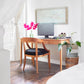 A minimalist home office setup showcases the American Shaker Writing Desk from Maple Corner Woodworks, crafted by Vermont artisans using sustainably harvested hardwood. The desk supports a computer monitor and keyboard, while pink flowers in a vase provide a splash of color. A world map decorates the wall, bathed in natural light streaming through the window.