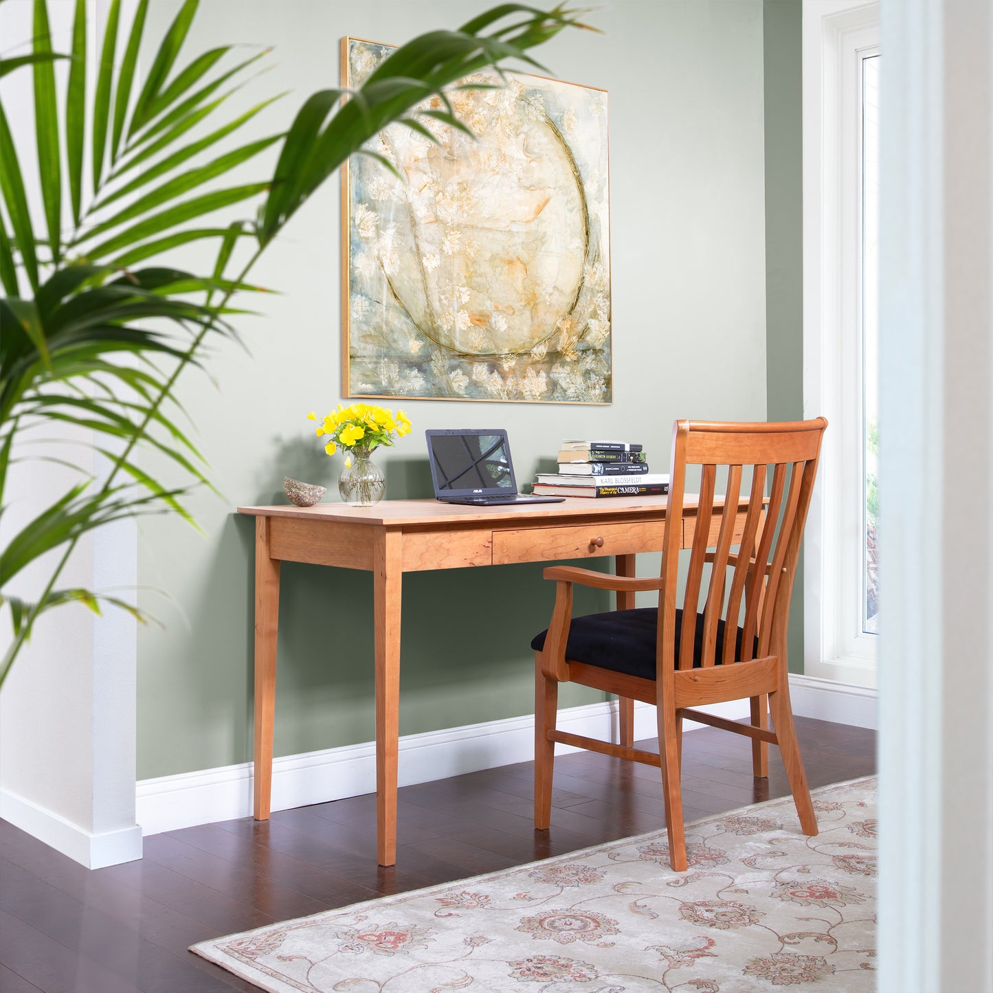 A cozy home office corner showcases an American Shaker Writing Desk from Maple Corner Woodworks, crafted from sustainably harvested hardwood by Vermont craftsmen. The desk supports a laptop, books, and a vase filled with yellow flowers. A large abstract painting graces the green wall as sunlight filters through the window onto a leafy plant.