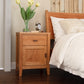 An Andover Modern 1-Drawer Nightstand with Door, crafted by Vermont woodworkers using sustainable hardwoods and sold by Maple Corner Woodworks, is placed next to a bed. On top of the nightstand, there are a vase with yellow tulips and