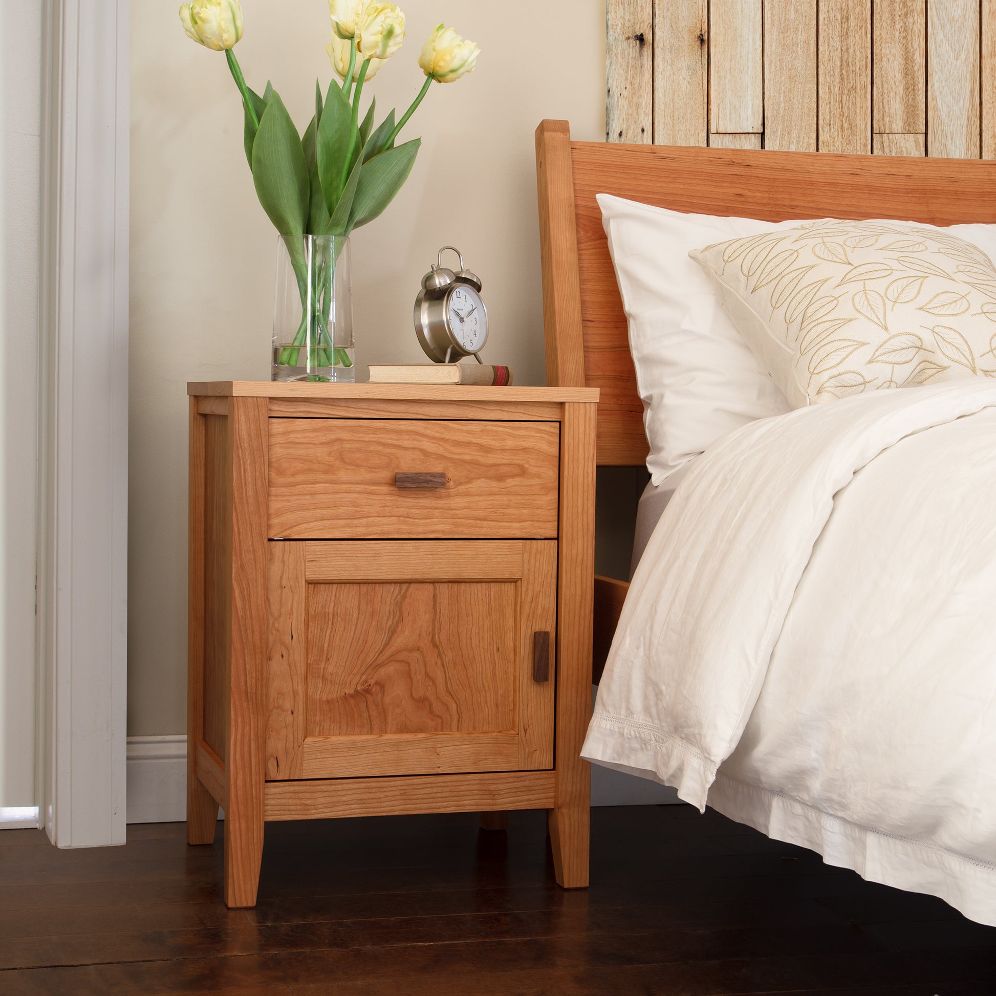 A Maple Corner Woodworks Andover Modern 1-Drawer Nightstand with Door, made of hardwood, placed next to a bed.