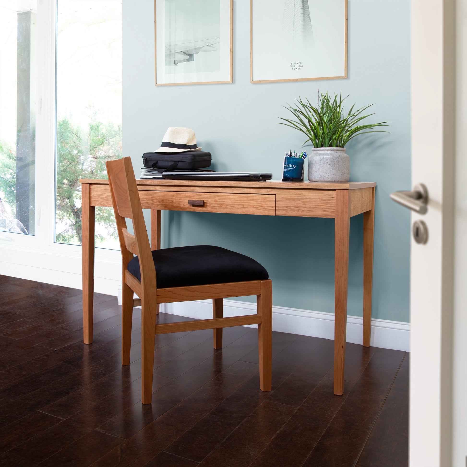 A Vermont Shaker Writing Desk by Maple Corner Woodworks, featuring a single drawer, is set against a light blue wall. Positioned in front of it, a wooden chair with a black cushion awaits. The desktop holds a plant, a stack of books, pens, and a hat beneath two framed artworks.