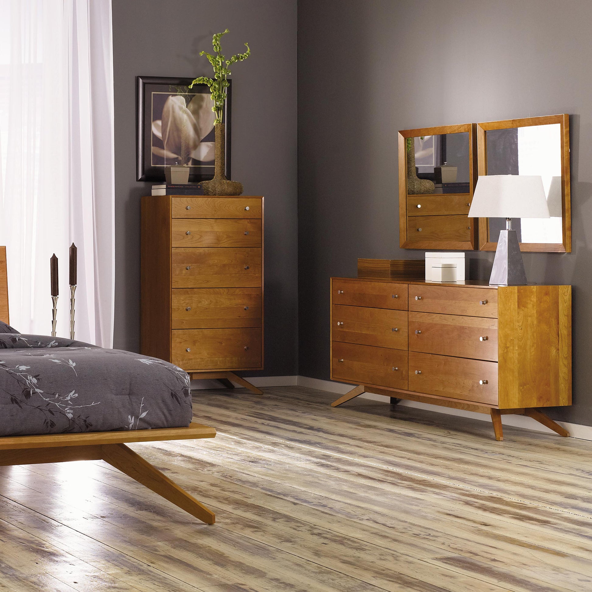 A modern American design bedroom features a wooden bed with a gray patterned comforter, handcrafted in Vermont. The room includes an Astrid 5-Drawer Chest by Copeland Furniture with a plant on top and another dresser with a mirror, lamp, and decorative items. It boasts gray walls, white curtains on large windows, and light wooden flooring.