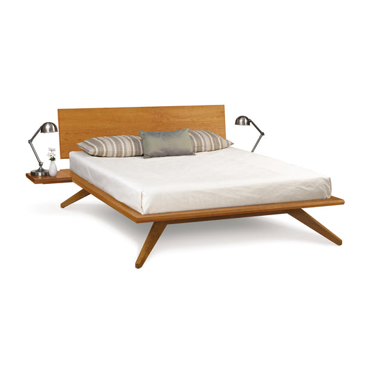 A modern Astrid Cherry platform bed crafted from American black walnut wood by Copeland Furniture, featuring angular legs and a simple headboard. The bed is dressed with a white comforter and gray striped pillows, flanked by two table lamps.