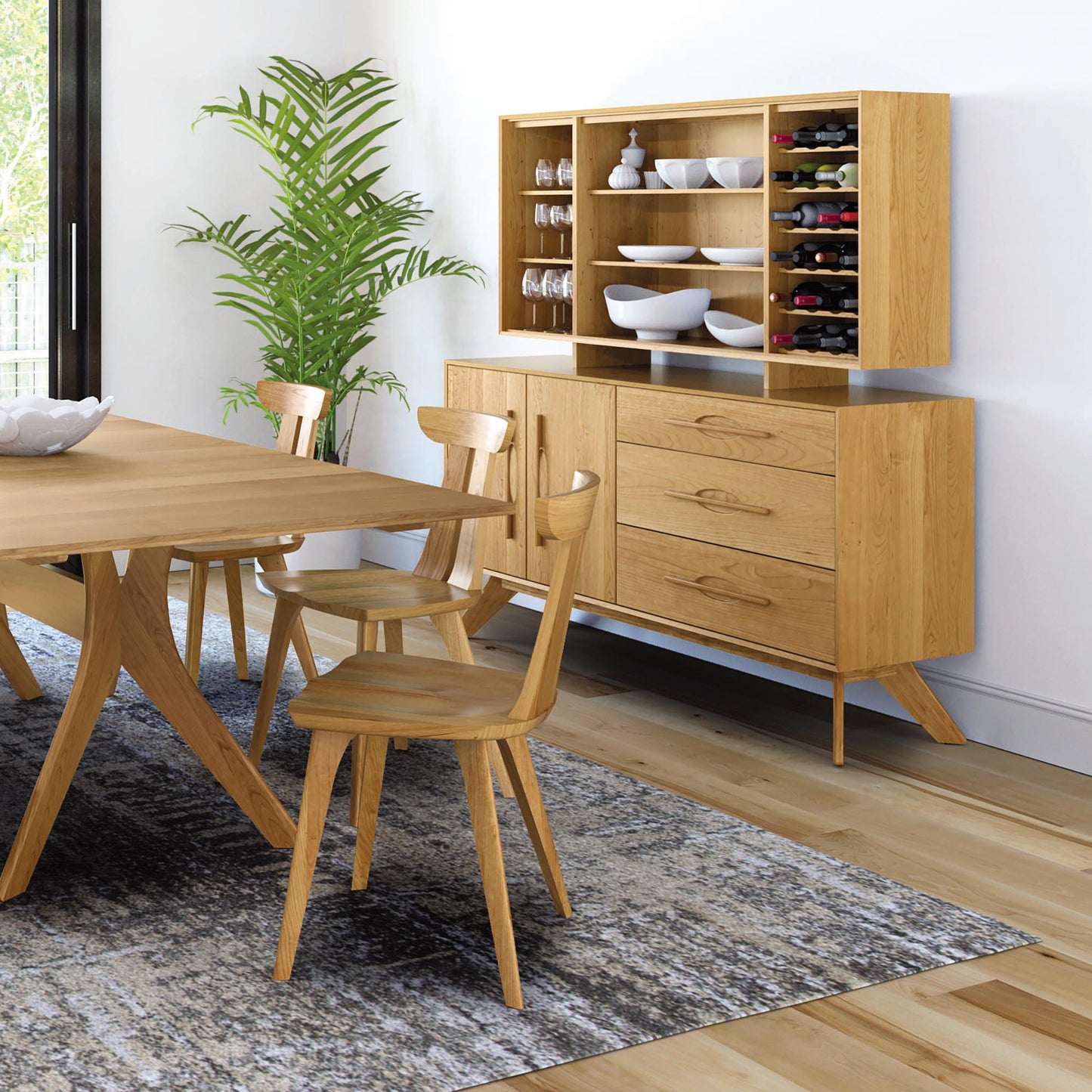 A modern dining room setup features a wooden dining table and four matching chairs on a textured rug. A handmade Audrey 2-Door 3-Drawer Buffet by Copeland Furniture, made from solid American hardwoods, holding dishes and wine bottles, stands against the wall beside a potted plant. Large windows provide natural light. Enjoy our lifetime guarantee.