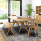 A modern dining room features the Audrey Extension Dining Table by Copeland Furniture, crafted from solid cherry wood, paired with matching chairs and a bench on one side. A white decorative bowl sits in the center of the table. A large potted plant stands near sliding glass doors, and a wooden cabinet with dishes is in the background.