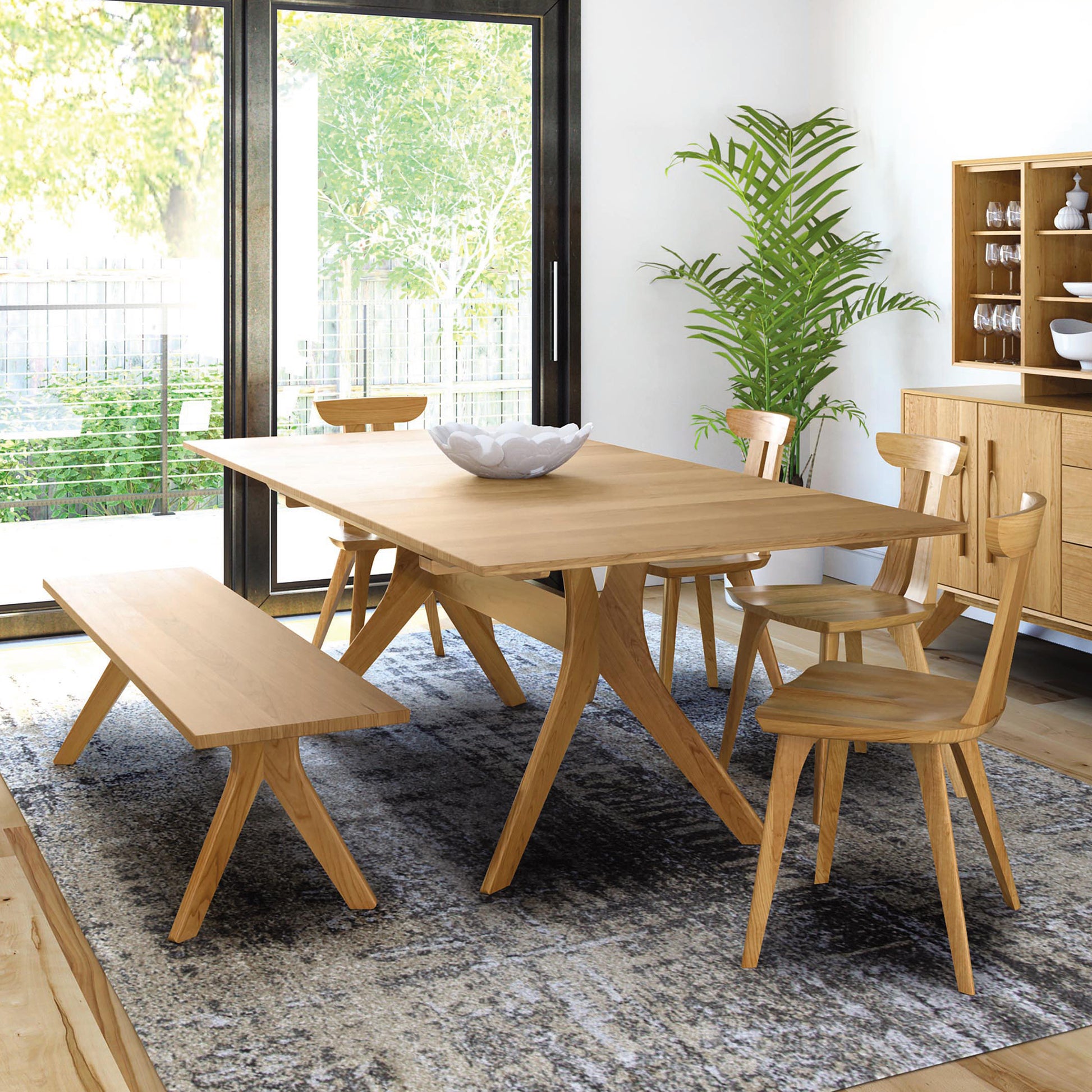 A modern dining room features the Audrey Extension Dining Table by Copeland Furniture, crafted from solid cherry wood, paired with matching chairs and a bench on one side. A white decorative bowl sits in the center of the table. A large potted plant stands near sliding glass doors, and a wooden cabinet with dishes is in the background.