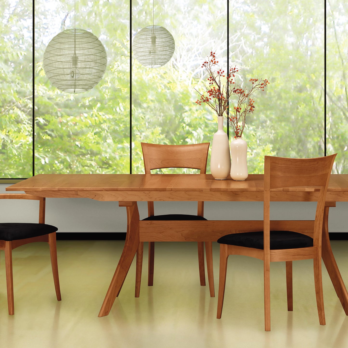 A solid Cherry wood dining table with three matching chairs and black cushioned seats is placed in front of large windows with a view of lush greenery. Two white pendant lights hang above the Copeland Furniture Audrey Extension Dining Table, which features a self-storing butterfly leaf mechanism.