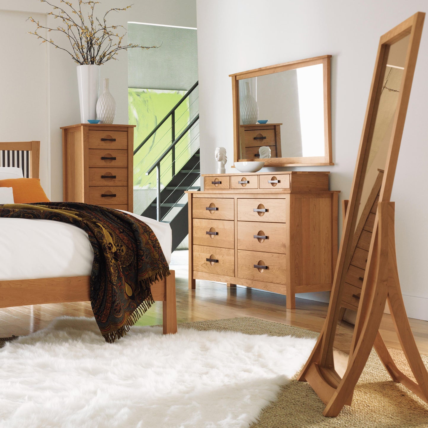 A bedroom with wooden furniture, featuring a bed, dresser with a mirror, Copeland Furniture's Berkeley 7-Drawer Lingerie Chest, and a full-length mirror on a stand. The bed is adorned with a white mattress and dark patterned blanket. A white rug covers part of the wooden floor, and a staircase can be seen in the background.