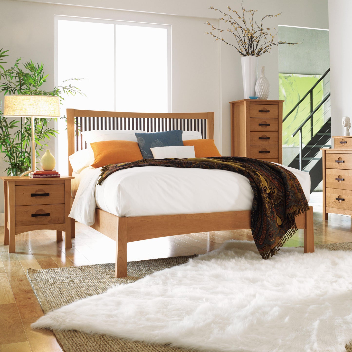 A modern bedroom with wooden furniture, including a Copeland Furniture Berkeley Cherry Platform Bed featuring white bedding and orange pillows, flanked by two wooden nightstands. Crafted from sustainably harvested hardwoods, a dresser and chest of drawers stand against the wall. A fluffy white rug covers part of the wooden floor.