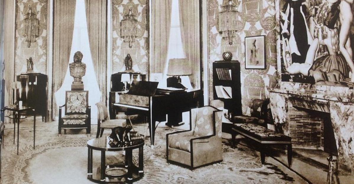 A sepia-toned photograph showcases an elegantly furnished room with tall windows draped in ornate curtains. The room features decorative chandeliers, various sculptures, a grand fireplace adorned with an intricate artwork, and classic furniture pieces.