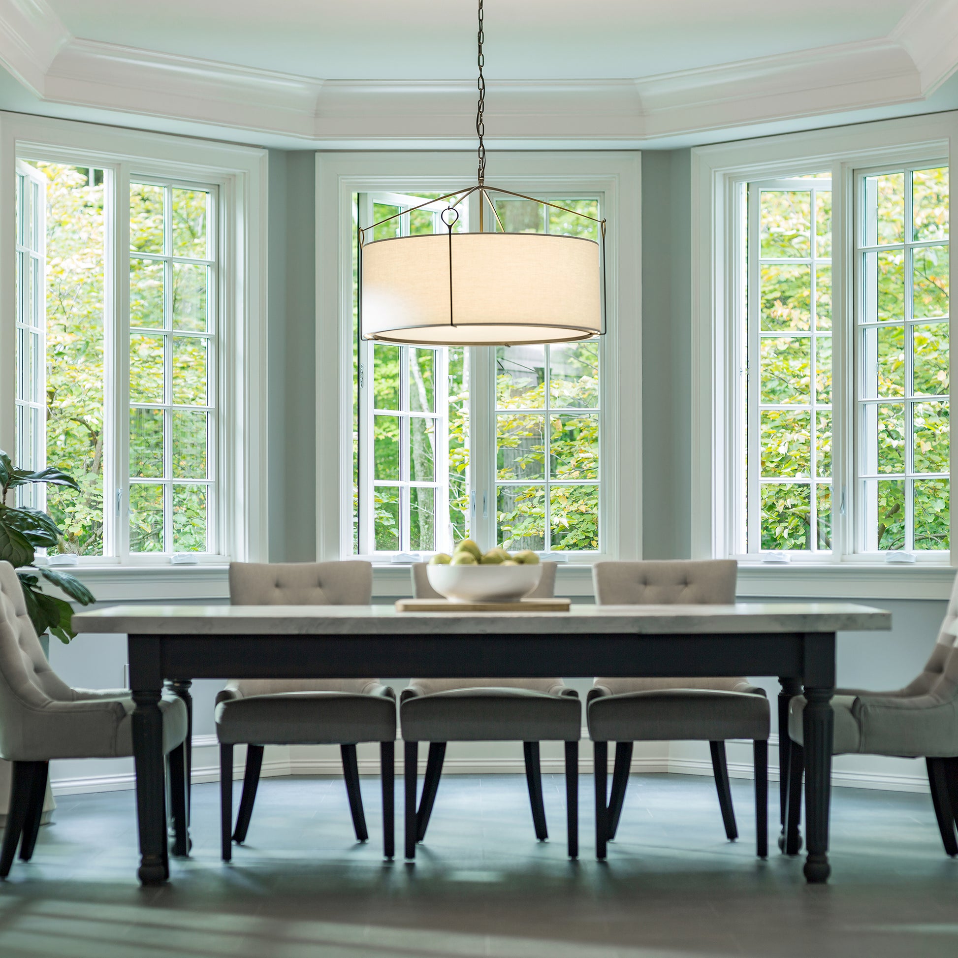 A bright and elegant dining room featuring a large wooden table with six chairs, a Hubbardton Forge Bow Large Pendant lamp above, and surrounded by tall windows showing lush greenery outside.
